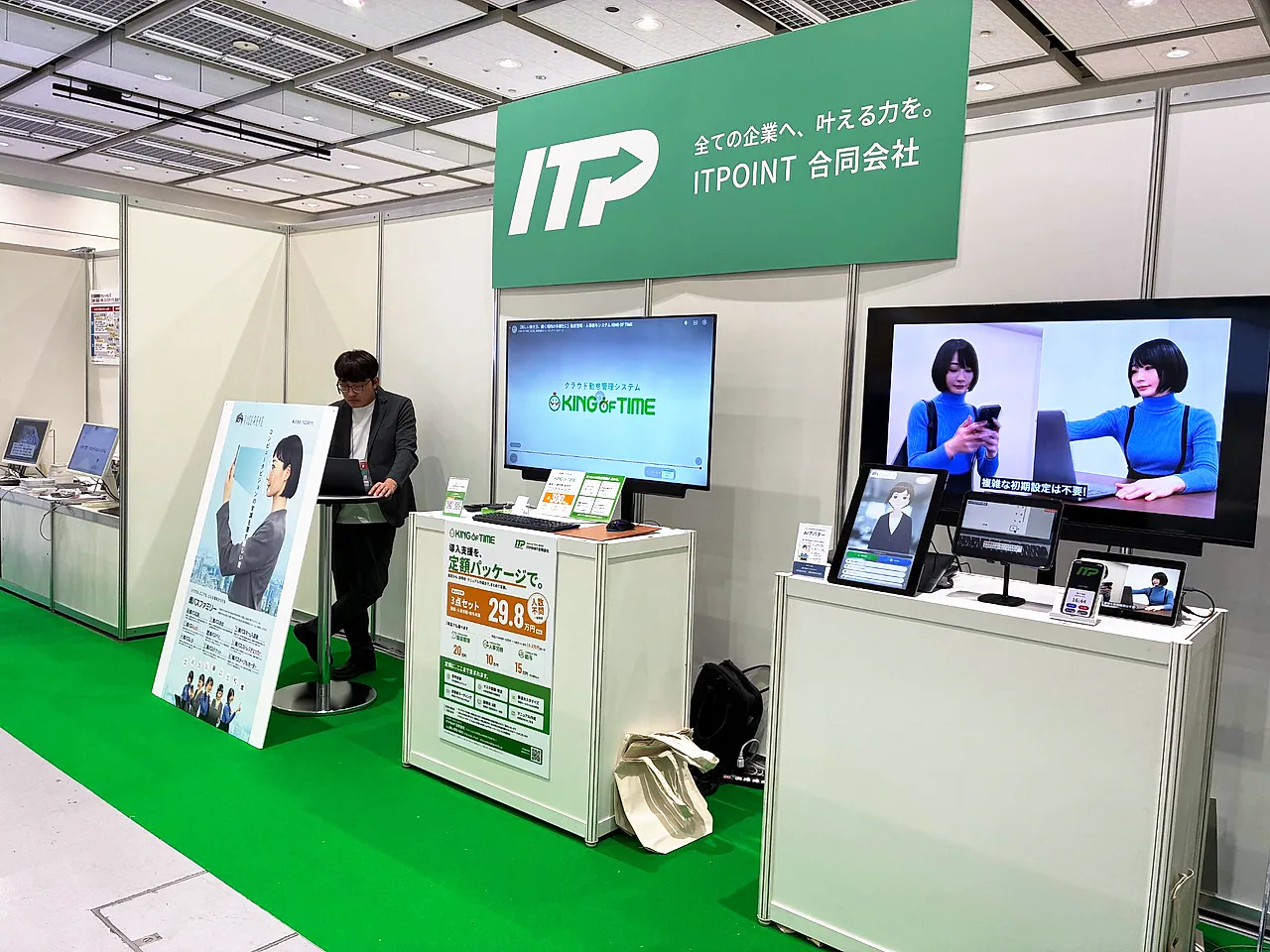 ブース全景。「全ての企業へ、叶える力を。」の看板の下に、KING OF TIMEのモニターと導入支援パッケージのポスター、AIアバターのタブレット、顔パスファミリーのポスターが並ぶ