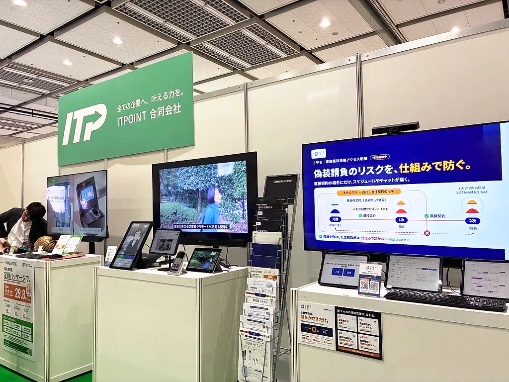 ケンセツミライのデモ。大型モニターに「偽装請負のリスクを、仕組みで防ぐ。」のスライド、手前にタブレットとパンフレットが並ぶ